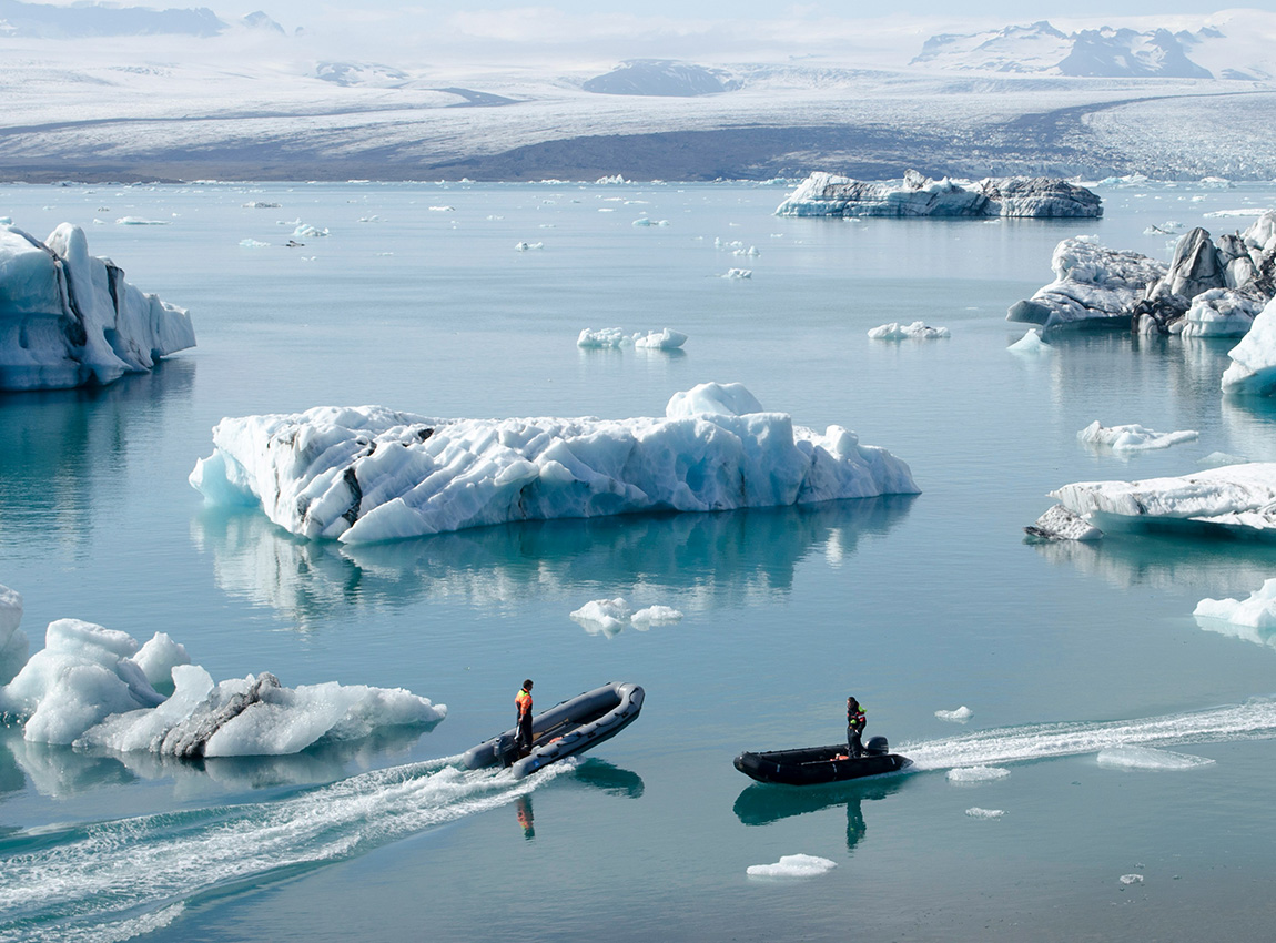 Two people in boats navigate between large icebergs on a calm, icy blue lake surrounded by snowy mountains.