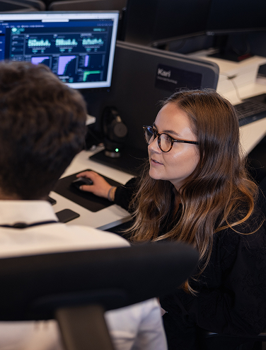 Two colleagues collaborating at a computer with data dashboards visible.