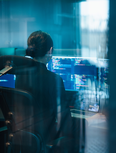 Person in an office chair looking at computer screens with code, seen through glass.