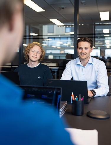 Three people sitting at a meeting table with laptops and office supplies in a modern office setting.