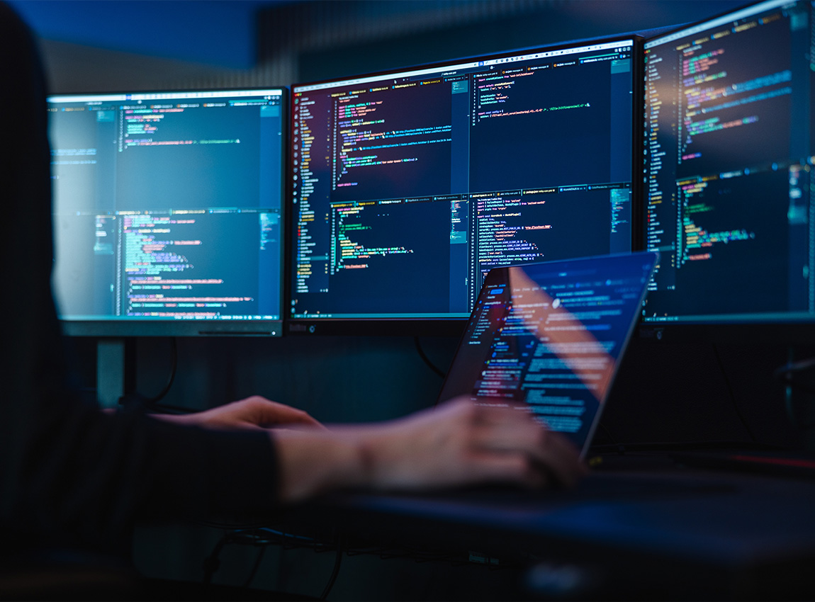 Desk with monitors showing code with the hands of a person typing on the keyboard.