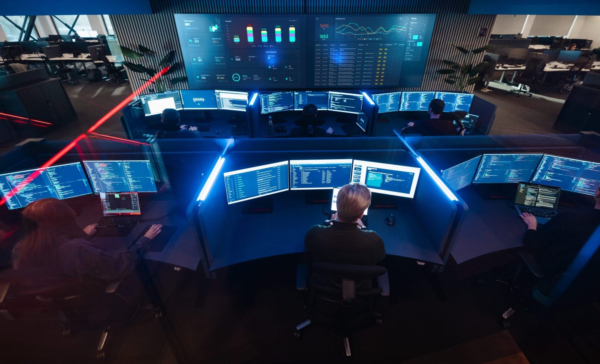 People monitoring multiple computer screens with data and code in a modern control room.